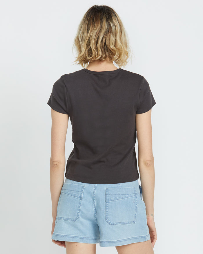 A woman with wavy, shoulder-length blonde hair faces away from the camera, wearing a Volcom Women's Have A Clue Tee and light blue denim shorts against a plain white background.