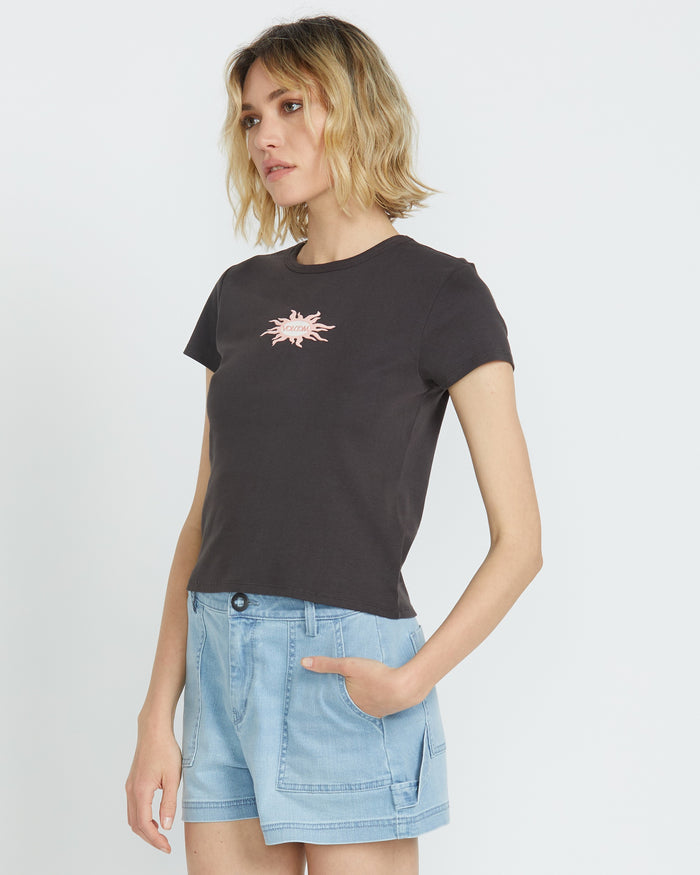 A woman with wavy blonde hair wears the Volcom Women's Have A Clue Tee, featuring a pink chest graphic, paired with light blue denim shorts. She stands against a plain white background, her left hand in her pocket.