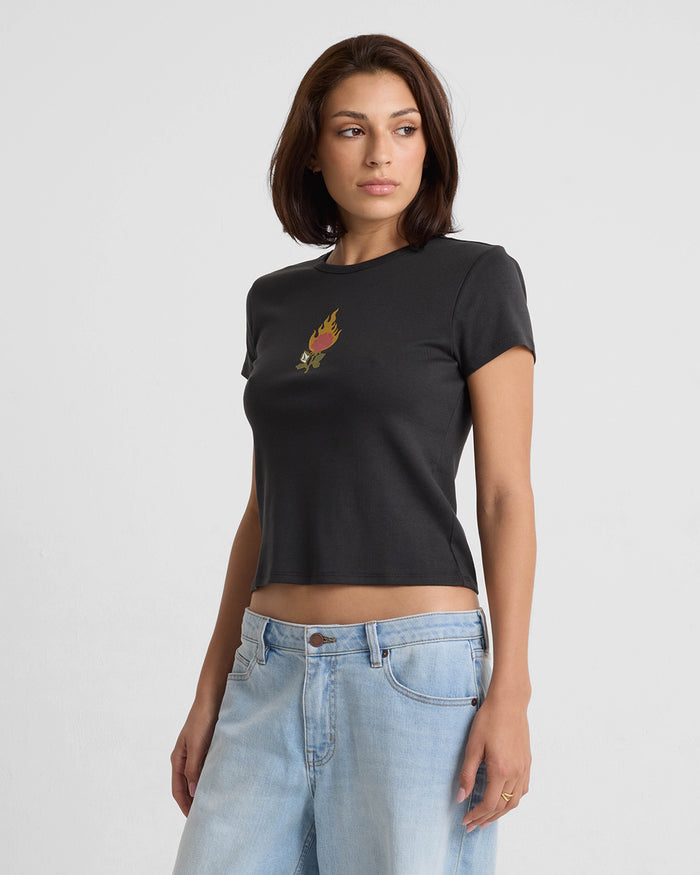 A woman with short brown hair models the Volcom Women's Have A Clue Tee, a fitted black T-shirt with a small flame graphic on the chest, paired with light blue jeans. She stands against a plain white background, looking slightly to her left.