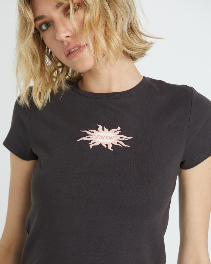 A woman with blonde hair models the Volcom Women's Have A Clue Tee, a black casual shirt with a pink abstract sunburst and "Volcom" on the front, looking to the side with a neutral expression.