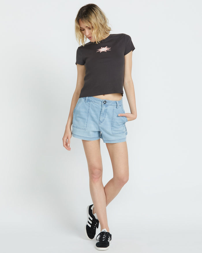 A person stands against a white background, wearing the Volcom Women's Have A Clue Tee in black, light blue denim shorts, and black sneakers with white stripes; one hand is in their pocket and their head is tilted down.