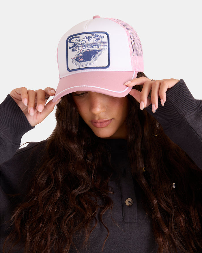A woman with long wavy brown hair in a black top wears the Volcom Women's Heartland Trucker, a pink-and-white mesh-back hat. She holds its brim with both hands.