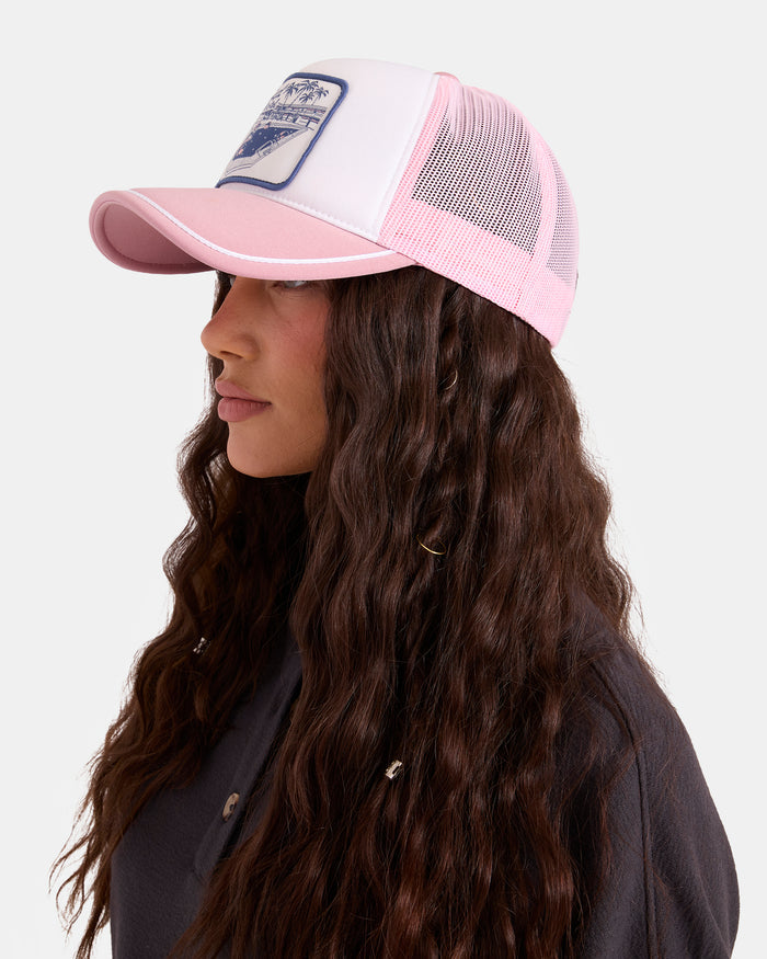 A woman with long, wavy brown hair models the Volcom Women's Heartland Trucker hat—a pink and white foam-front cap with a front patch and mesh back—paired with a dark shirt, shown in profile against a plain white background.