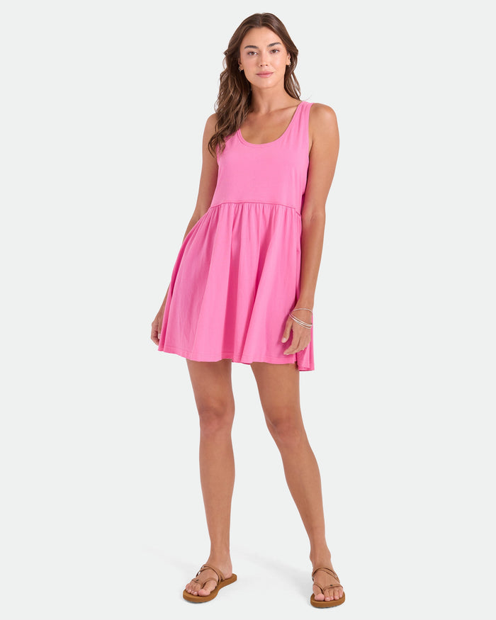 A woman poses against a white background wearing the Volcom Women's Hi Lo Tide Dress, a pink sleeveless mini dress made from 100% cotton, paired with tan sandals and simple jewelry. One hand rests on her dress.