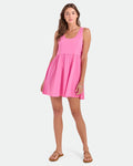 A woman poses against a white background wearing the Volcom Women's Hi Lo Tide Dress, a pink sleeveless mini dress made from 100% cotton, paired with tan sandals and simple jewelry. One hand rests on her dress.