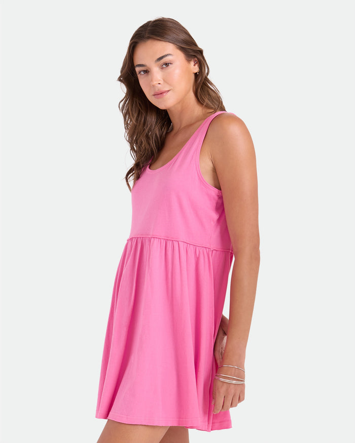 A woman with long brown hair wears the Volcom Women's Hi Lo Tide Dress, a sleeveless, scoop-neck pink mini dress, paired with silver bangles, standing against a plain light background.