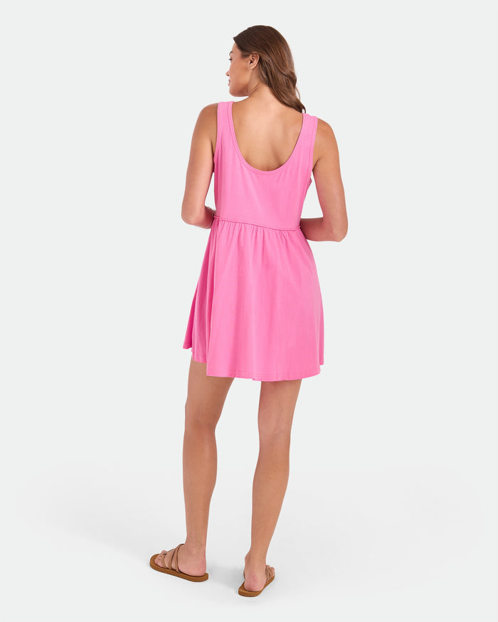 A woman with light skin and long brown hair faces away, wearing the Volcom Women's Hi Lo Tide Dress in pink (100% cotton, sleeveless, scoop-back) and brown sandals. The background is plain white.