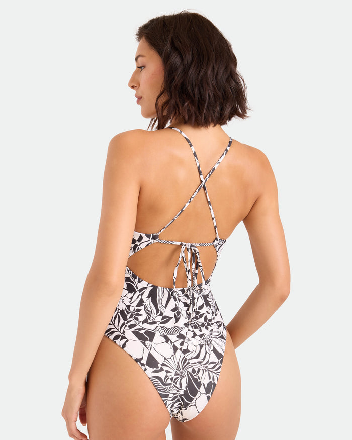 A woman with short brown hair models the Volcom Women's Hibiscus Diamond Keyhole One-Piece, featuring black and white florals, crisscrossed adjustable straps, and a tie-back, standing against a plain light background facing away.