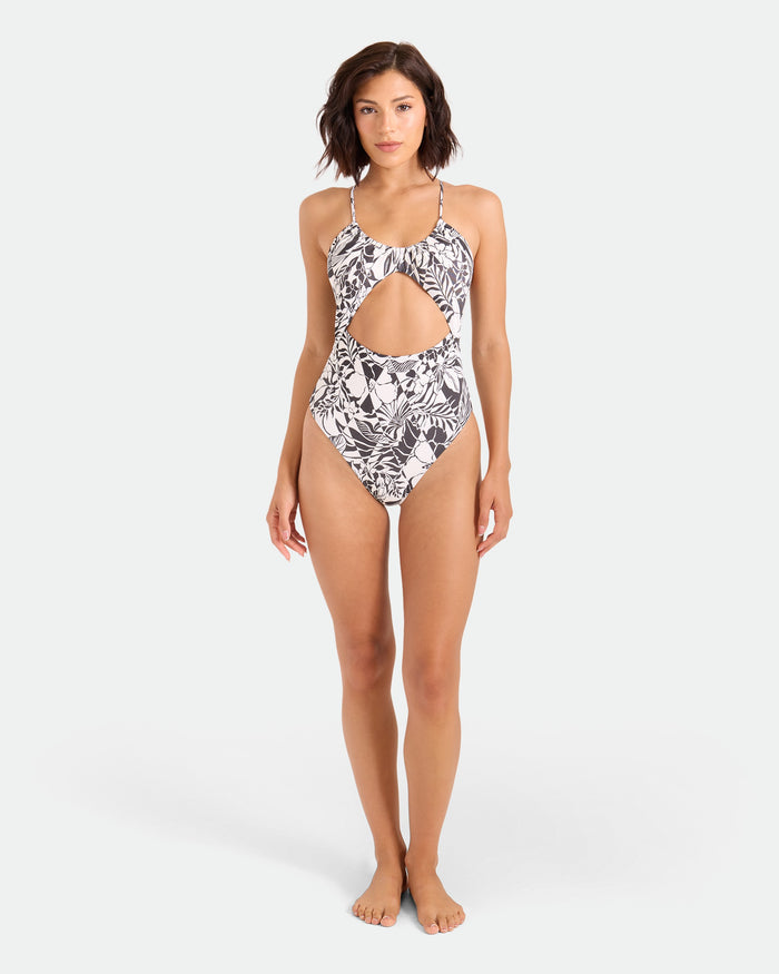 A woman stands barefoot in Volcom's Women's Hibiscus Diamond Keyhole One-Piece, a black and white recycled nylon swimsuit with a halter neck, abstract leaf pattern, and cutout below the chest, facing forward against a plain light background.