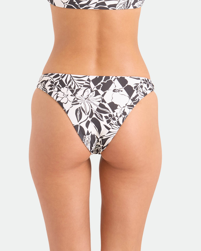 A person is shown from the waist down wearing Volcom's Women's Hibiscus Diamond Midrise Skimpy Reversible Bikini Bottoms, featuring a black and white floral print, made from recycled nylon swimwear, against a plain light background.