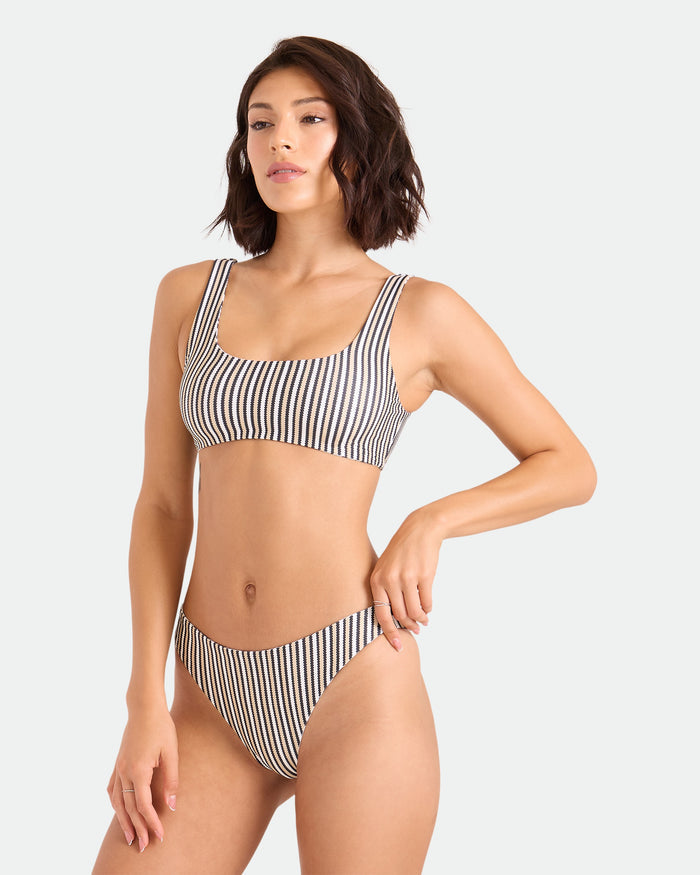 A woman with short dark hair wears the Volcom Women's Hibiscus Diamond Midrise Skimpy Reversible Bikini Bottom, standing with one hand on her hip against a plain light background.