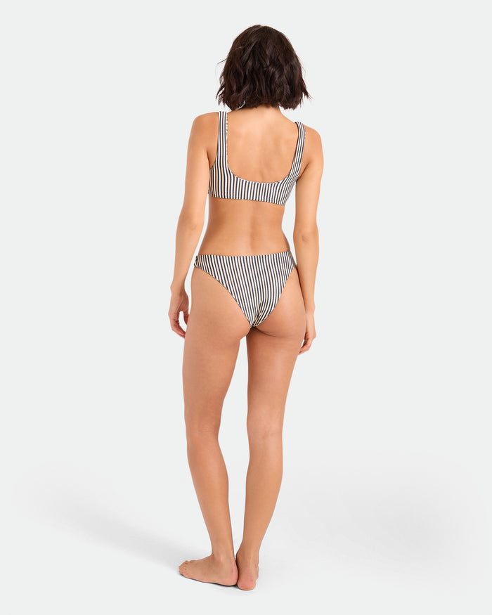 A woman stands barefoot facing away, wearing Volcom's Women's Hibiscus Diamond Midrise Skimpy Reversible Bikini Bottom, against a plain light background.
