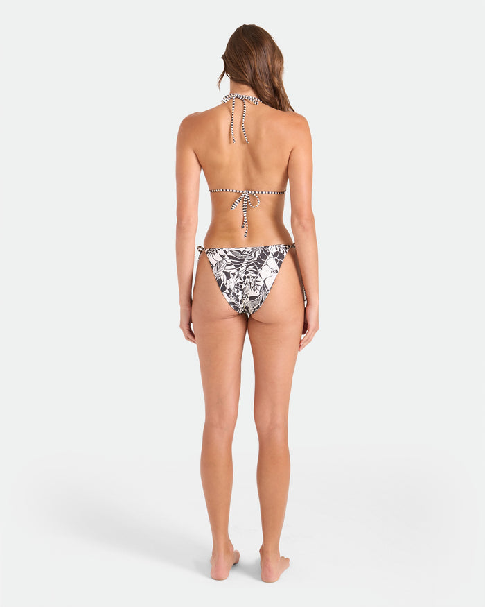 A woman stands barefoot facing away, wearing the Volcom Women's Hibiscus Diamond Reversible Triangle Top with a striped design and black-and-white floral bikini bottoms, set against a plain white background.