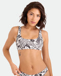 A woman with short brown hair wears the Volcom Women's Hibiscus Diamond Scoop Bralette bikini, featuring black and white hues and adjustable straps, made from recycled nylon. She stands against a plain light background, her butterfly tattoo showing.