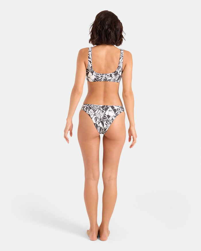 A woman with short brown hair stands barefoot, facing away, wearing the Volcom Women's Hibiscus Diamond Scoop Bralette with adjustable straps and a black and white floral bikini made from recycled nylon against a white background.
