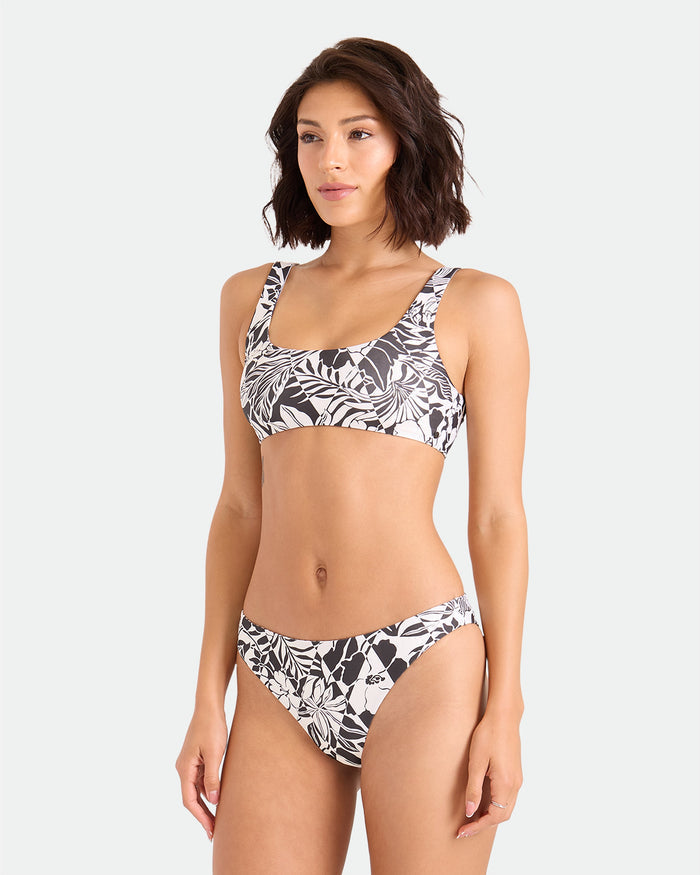 A woman models the Volcom Women's Hibiscus Diamond Scoop Bralette in black and white floral, featuring adjustable straps and made from recycled nylon, standing against a plain light background with a neutral expression.