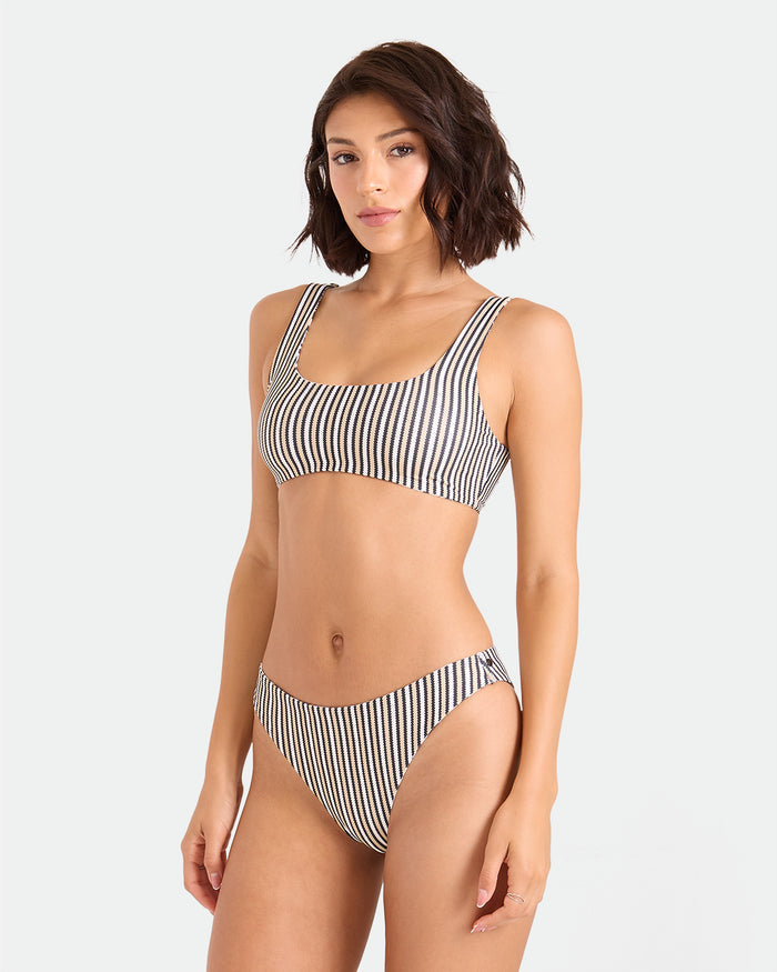 A woman with shoulder-length brown hair models the Volcom Women's Hibiscus Diamond Scoop Bralette, standing against a plain light background and looking into the camera.