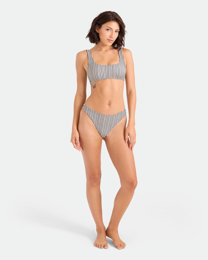 A woman with short brown hair and a neutral expression stands barefoot against a plain background, wearing the Volcom Women's Hibiscus Diamond Scoop Bralette—featuring black-and-white vertical stripes, adjustable straps, and recycled nylon fabric.