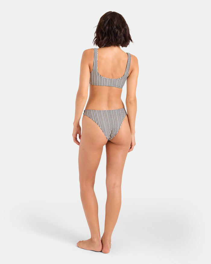 A woman with chin-length wavy hair stands barefoot on a plain white background, facing away. She is wearing Volcom's Women's Hibiscus Diamond Scoop Bralette, featuring black and white vertical stripes and adjustable straps made from recycled nylon.