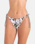 A woman wears Volcom’s Women's Hibiscus Diamond Tie Side Full Reversible Bikini Bottom, shown waist to mid-thigh against a light background. Features black and white floral print, tie sides, and eco-friendly recycled nylon.
