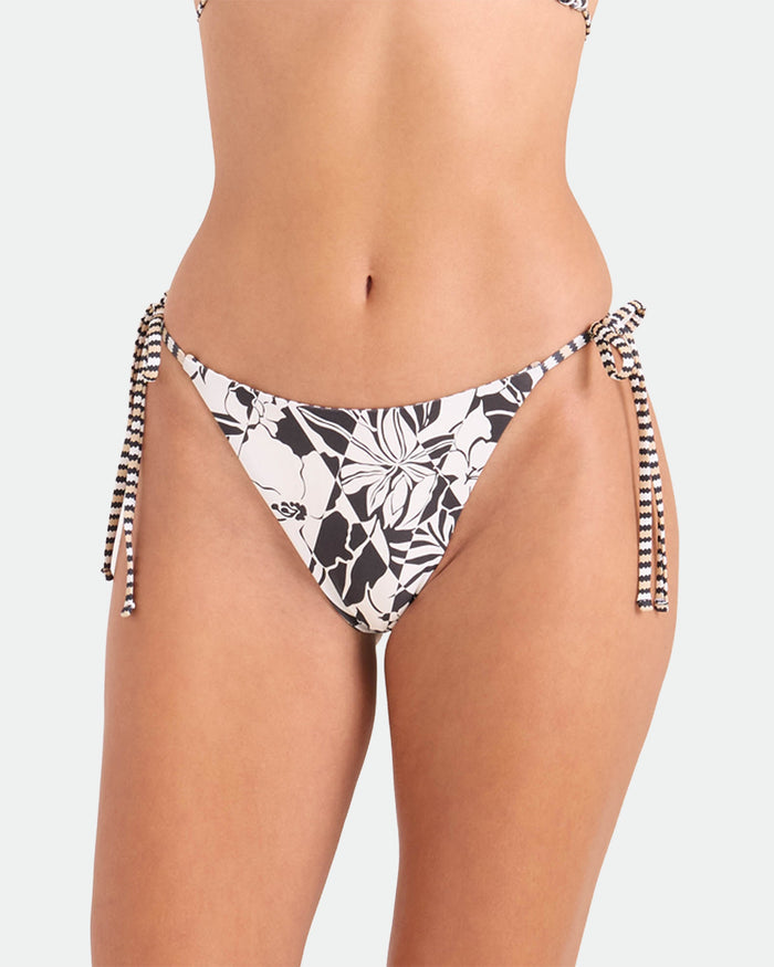 A woman wears Volcom’s Women's Hibiscus Diamond Tie Side Full Reversible Bikini Bottom, shown waist to mid-thigh against a light background. Features black and white floral print, tie sides, and eco-friendly recycled nylon.