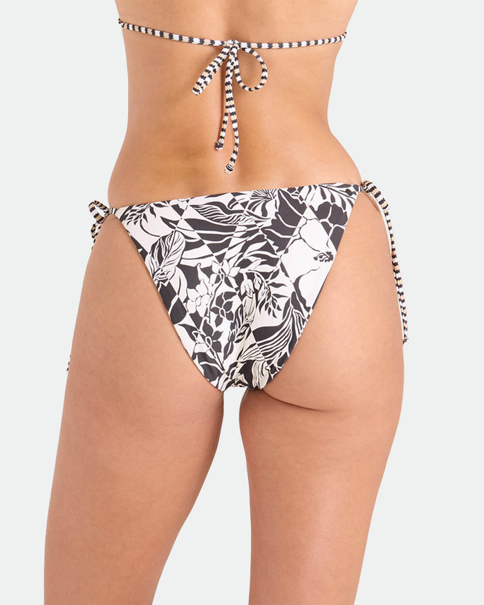 A woman is shown from the back wearing Volcom's Women's Hibiscus Diamond Tie Side Full Reversible Bikini Bottom in a black and white tropical leaf print with side ties, paired with a matching bikini top, set against a plain light background.