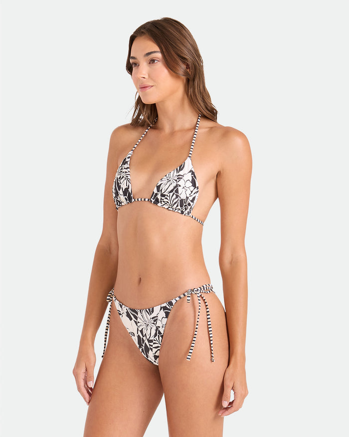 A woman wears the Volcom Women's Hibiscus Diamond Tie Side Full Reversible Bikini Bottom, featuring a black and white floral print with tie-side details, crafted from recycled nylon. She stands slightly sideways against a plain background, arms relaxed.