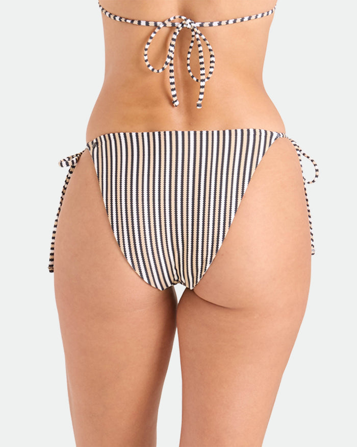 A woman is shown from the back wearing Volcom's Women's Hibiscus Diamond Tie Side Full Reversible Bikini Bottom, featuring vertical black and white stripes, made from recycled nylon, against a plain white background.
