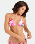 A woman with short brown hair wears the Volcom Women’s In A Daze Triangle Top, a colorful floral bikini featuring adjustable tie straps, and looks down as she adjusts the side tie of her bikini bottom against a plain white background.