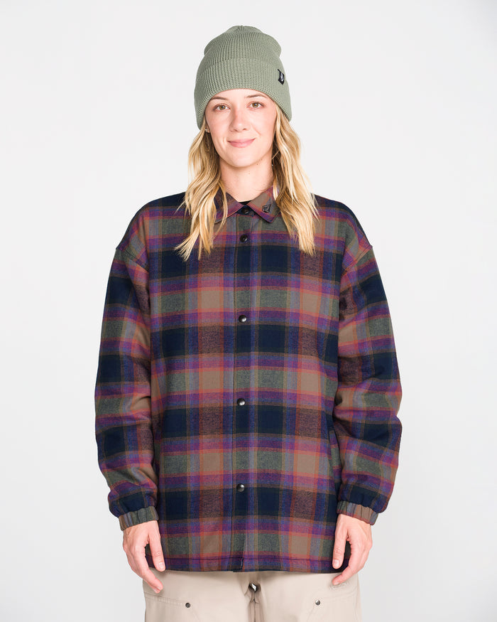 A person stands facing forward against a plain white background, hands relaxed by their sides, wearing the Volcom Women’s Insulated Riding Flannel Jacket—a plaid flannel jacket with taffeta lining—and a green knit beanie.