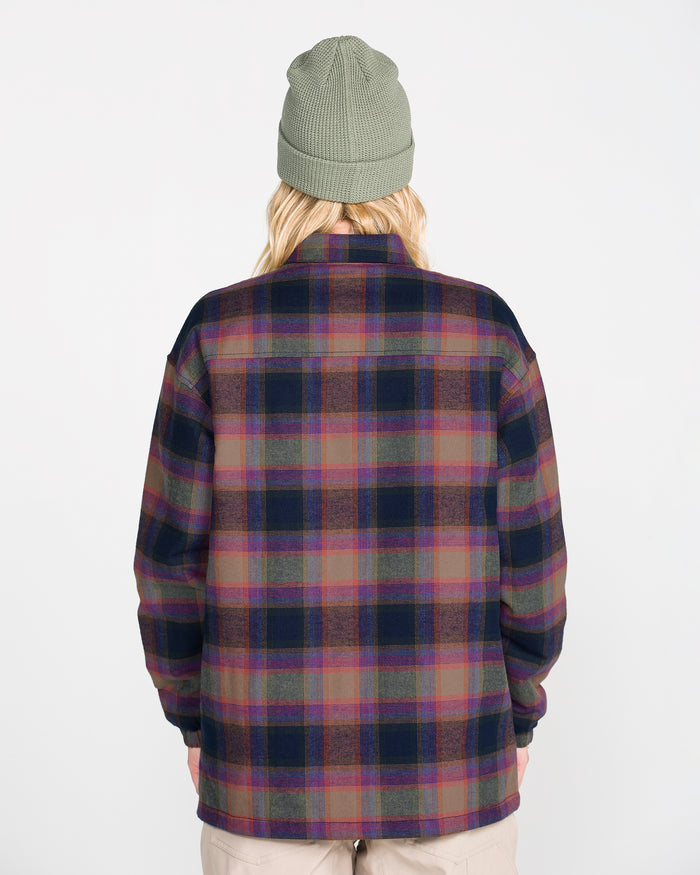 A person is shown from behind, wearing a green knit beanie and the Volcom Women’s Insulated Riding Flannel Jacket in a plaid pattern with shades of purple, pink, and green. The jacket features a soft taffeta lining for added comfort. The background is plain white.