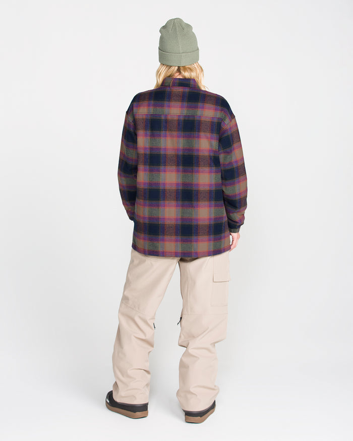A person is seen from behind, standing against a plain white background, wearing the Volcom Women’s Insulated Riding Flannel Jacket in dark tones with taffeta lining, paired with a green beanie and loose beige pants featuring pockets.