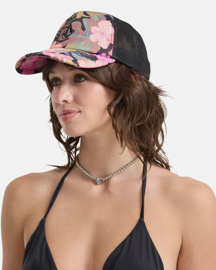 A woman in a black bikini top, silver heart necklace, and the Volcom Women's Into Paradise Hat with a floral pattern and mesh back stands against a plain white background, facing slightly to the side.