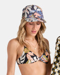 A woman wearing the Volcom Women's Into Paradise Trucker Hat looks down with a slight smile. She has loose, wavy blonde hair and layered gold necklaces, pairing the hat with a floral bikini top for a relaxed, beach-ready style.