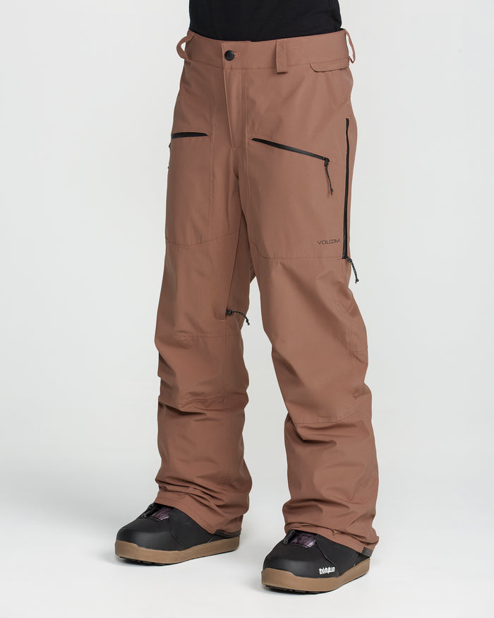 A person is wearing Volcom Women’s Karat Stretch 30K Pants in brown, featuring black zippers and paired with black boots, standing against a plain white background. Only the lower half of their body is visible.