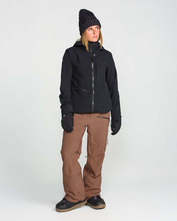 A person stands against a plain white background, looking to the side, wearing a black beanie, black jacket, black gloves, and loose brown Volcom Women’s Karat Stretch 30K Pants.
