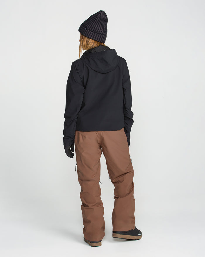 A person stands facing away against a plain white background, wearing a black jacket, black gloves, a dark beanie, and Volcom Women’s Karat Stretch 30K Pants—women’s waterproof ski pants crafted from recycled polyester.