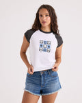 A woman wears Volcom's Women's Kick It Game Day Crop Tee—a white T-shirt with vintage black sleeves and a blue geometric design on the chest—paired with denim shorts, standing against a plain white background.