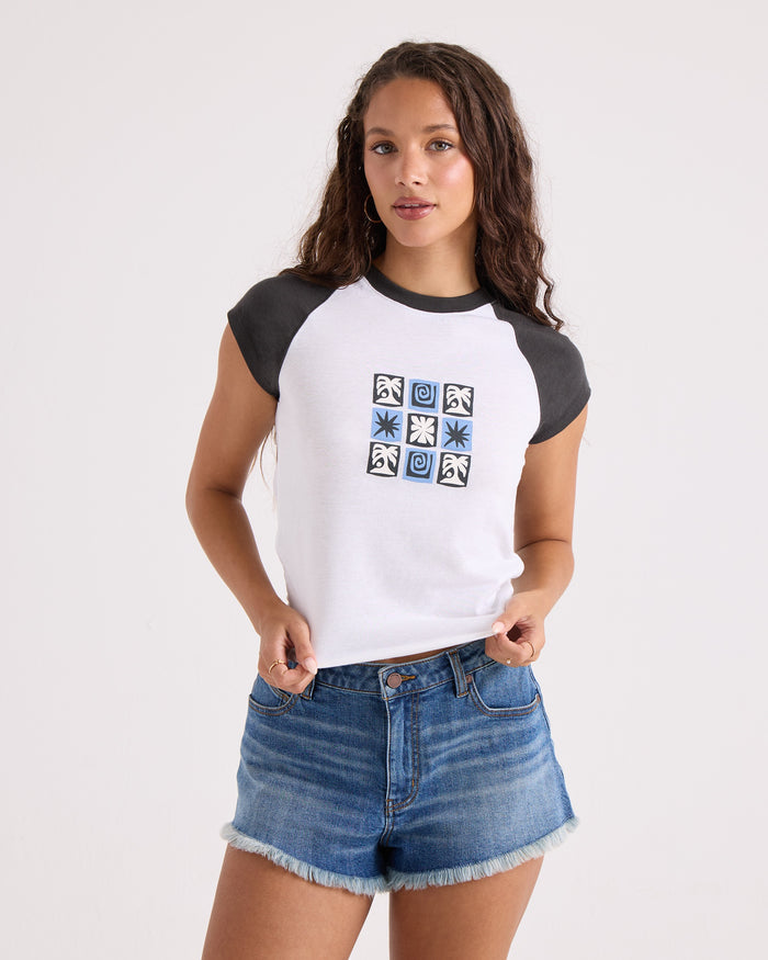A woman wears Volcom's Women's Kick It Game Day Crop Tee—a white T-shirt with vintage black sleeves and a blue geometric design on the chest—paired with denim shorts, standing against a plain white background.