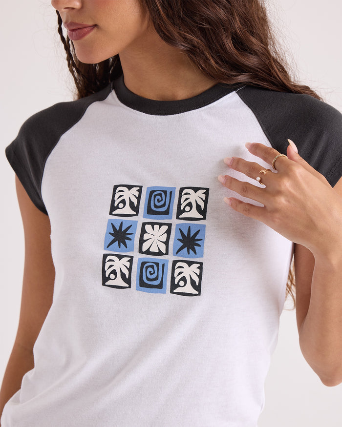 A person wearing the Volcom Women's Kick It Game Day Crop Tee—a white and black raglan shirt featuring a blue and black geometric pattern of squares and swirls on the front, reminiscent of vintage sportswear—touches their shirt with one hand.
