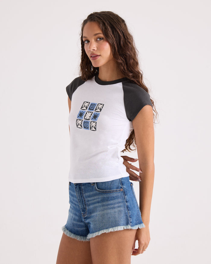 A woman with long curly hair stands against a plain white background, looking at the camera with one hand by her side. She is wearing the Volcom Women's Kick It Game Day Crop Tee—a white and vintage black raglan t-shirt with a graphic—paired with blue denim shorts.