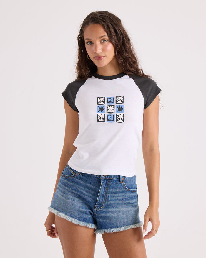 A young woman with long wavy hair wears the Volcom Women's Kick It Game Day Crop Tee, featuring white and vintage black colors with geometric designs, paired with frayed denim shorts as she stands against a plain light background.