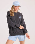 A woman wearing a light blue baseball cap, a Volcom Women's Levy Crew Fleece sweatshirt in soft cotton-blend with a small chest graphic, and light blue denim shorts poses against a plain light background.