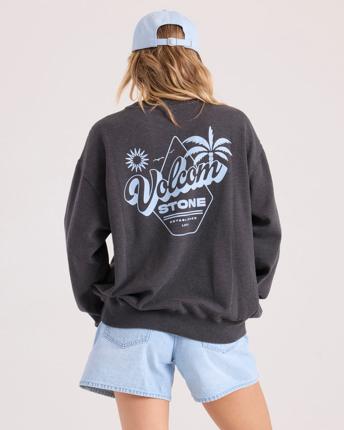 A person stands with their back to the camera, wearing a light blue cap, Volcom Women's Levy Crew Fleece with a tropical graphic on the back, and light denim shorts against a plain background.