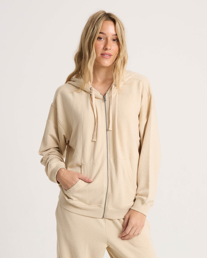 A woman with long blonde hair wears the Volcom Women's Lil Frenchie Zip Hoodie, standing against a plain white background, facing forward with one hand in her pocket.