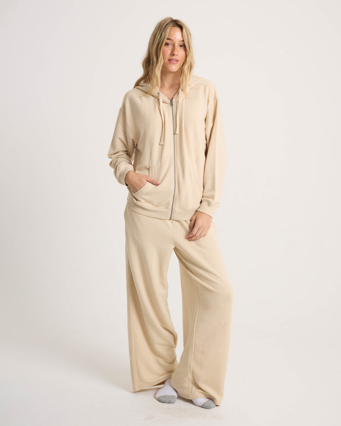 A woman with long blonde hair stands against a plain background in the Volcom Women's Lil Frenchie Zip Hoodie and matching beige wide-leg lounge pants, her hand in one pocket, wearing white socks.