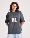 A woman stands against a plain background, hands behind her back and looking at the camera, wearing light blue jeans and the Volcom Women's Line Up Base Tee—a vintage black oversized t-shirt with "VOLCOM 91" in distressed white letters.