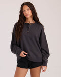 A young woman with long, wavy brown hair wears the Volcom Women's Lived In Fleece Henley and black denim shorts, standing against a plain light background and looking at the camera.