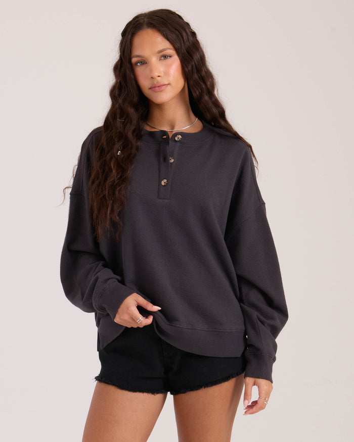 A woman with long wavy brown hair wears the Volcom Women's Lived In Fleece Henley and black shorts. She stands against a plain light background, looking at the camera with a relaxed expression.
