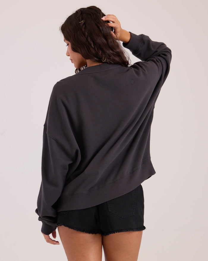 A woman stands with her back to the camera, touching her hair, wearing the Volcom Women's Lived In Fleece Henley and black denim shorts.